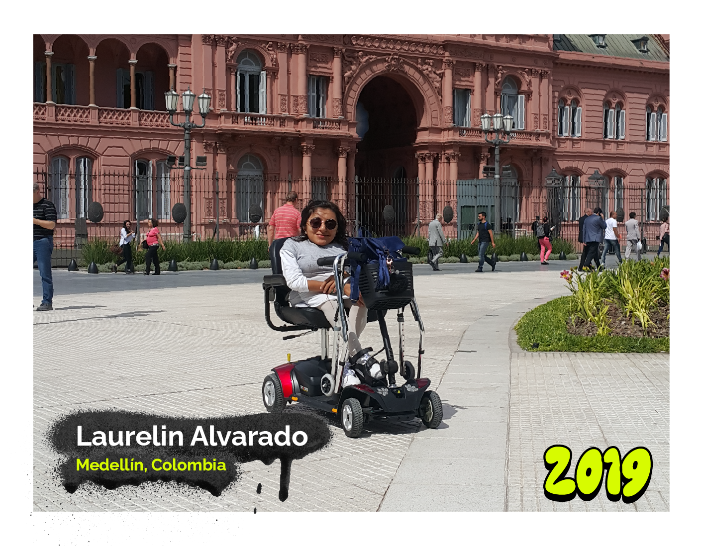 Fotografía de una mujer llamada Laurelin Alvarado, de Medellín, Colombia, está sentada en una silla de ruedas eléctrica frente a un edificio histórico de color rosado con arquitectura clásica. La imagen tiene un marco con detalles en colores vivos y el año '2019' destacado en la esquina inferior derecha.