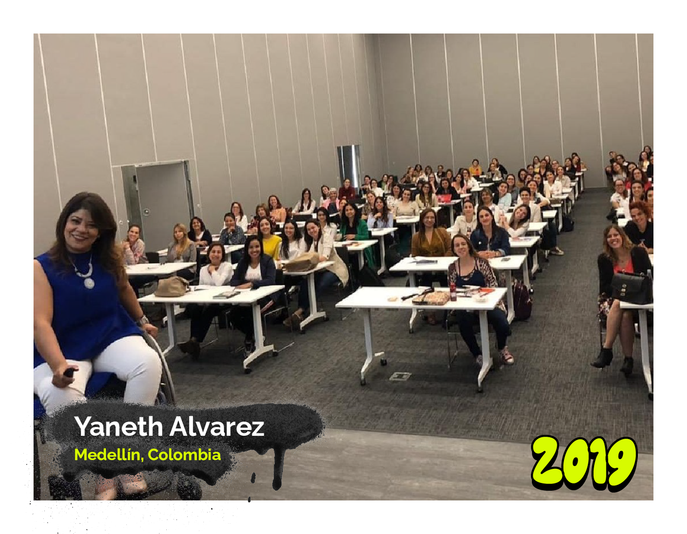 Fotografía de Yaneth Álvarez, de Medellín, Colombia, está sentada en una silla de ruedas frente a un auditorio lleno de personas, en su mayoría mujeres, en un salón amplio de paredes grises. Lleva una blusa azul sin mangas, un pantalón blanco y un collar largo, mientras sonríe cálidamente a la audiencia. En la esquina inferior derecha se destaca el año '2019' con un diseño colorido.